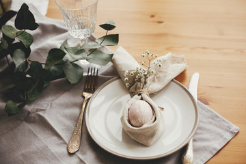 Stylish Easter brunch table setting with egg in easter bunny napkin on  table. Modern natural dyed pink easter egg on napkin with bunny ears, flowers on plate and cutlery. Easter table decorations.