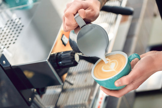 Coffee Passion. Young barista at coffee shop pouring steamed milk into cup making latte art heart picture close-up