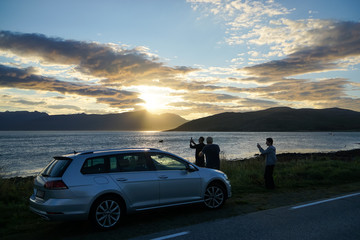 Fototapeta na wymiar Three men having a stop to photograph sunset on road trip at Arctic Senja Tromso Norway beautiful fjord car standing by
