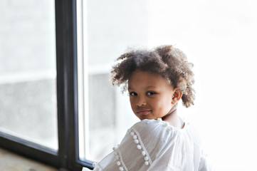 Close-up portrait of cute little african american kid girl in a white dress. She's posing sitting by the window.  Studio shoot. .