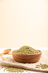 Wooden bowl with raw green mung bean and wooden spoon on a white and orange background. Side view, selective focus, copy space.