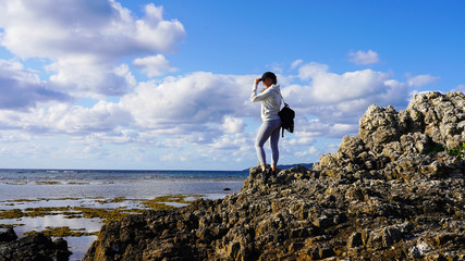 Obraz premium Young girl traveler with a backpack on her shoulders stands on top of a rocky mountain on the Pacific Ocean at low tide. Sea view on the Japanese island of Ishigaki in Okinawa. Background of clear sky