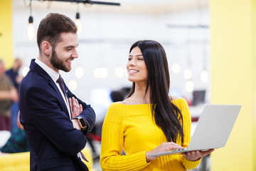 Beautiful Caucasian dark haired girl in yellow posing with businessman wearing suit. Happy colleagues with a laptop in a modern office