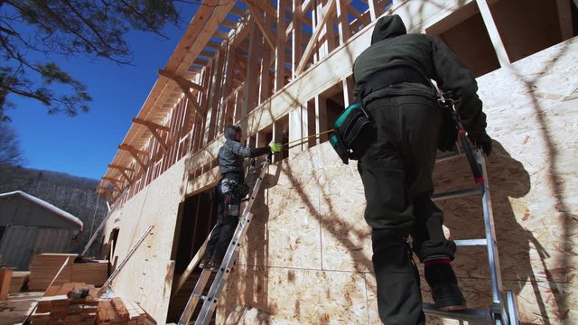 Two builders on ladders are masuring plywoods with measuring tape. Frame house building site