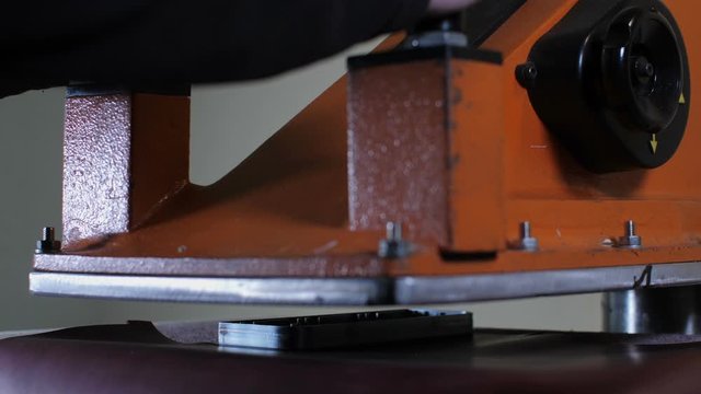 Close-up of craftsman's hands cutting out leather pattern using sharp metal mold and pressing machine while working in leather crafting atelier. Leatherworker in process of exclusive manufacturing