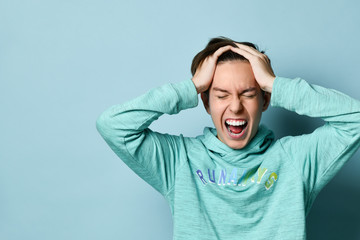 Half-length emotional portrait of a Caucasian teenage boy in a blue hoodie. Excited guy screaming holding his head in his hands