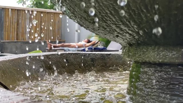 slow motion reveal of a young woman suntanning. Beautifully framed by a fountain and drops of water. Bali holidays concept.