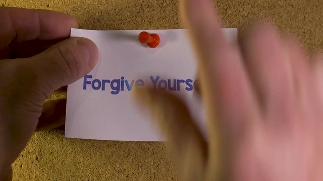Man hands pin a note on an office pinboard saying "Forgive Yourself".