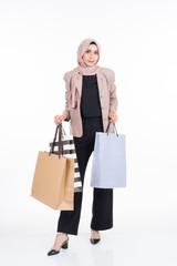 A beautiful Asian Muslim woman wearing officewear and hijab poses with shopping bags isolated on white background. Full length portrait for business and commerce concepts.