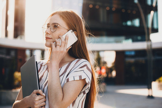 Charming Business Woman With Red Hair And Eyeglasses Talking On Phone During A Walk Outside The Office With Her Computer