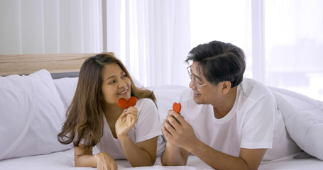 Happy asian couple holding red heart decorations together in a bedroom.