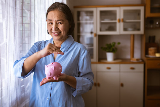 Asain Elder  Putting Coin Money To Piggy Bank Saving, Family, Savings, Age And People Concept - Smiling Senior Couple With Money And Piggy Bank At Home.