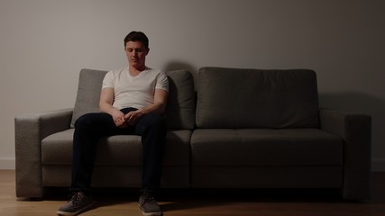 Lone young adult man sits on a sofa at home in the night