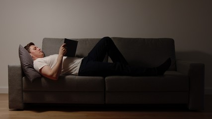 Young adult man lies on a sofa and reads a book in the evening - 327737334