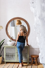 A girl of European appearance is standing near a table with a mirror and prenadlezhnostyami for makeup, perfume, sink, dressed in jeans