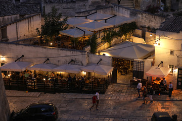 Obraz premium Evening view of the city of Matera, Italy, with the colorful lights highlighting patios of sidewalk cafes in the Sassi di Matera a historic district in the city of Matera. Basilicata. Italy