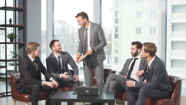 Young caucasian business colleagues sit on leather chairs and discuss together in modern office, wearing elegant suits. big panoramic window in the background