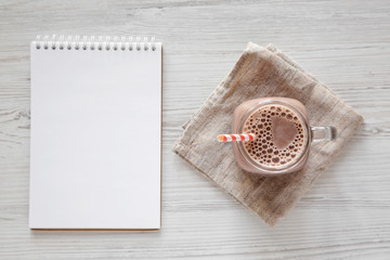Homemade New England Chocolate Milkshake in a Glass Jar Mug, blank notepad on a white wooden background, top view. Overhead, from above.