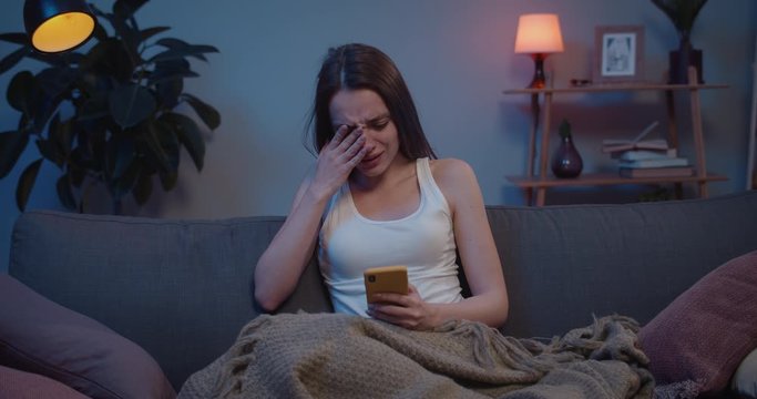 Front view of girl sitting on sofa with smartphone and thinking about sudden problems. Young sad woman covering face with hand and crying after reading message with bad news.