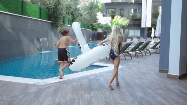 girl and boy run by the pool with pool toy in their hands.