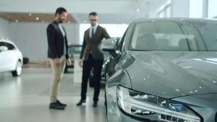 Handsome guy is talking to professional sales agent in car dealership, focus on expensive automobile. Transportation, luxury and modern vehicles concept. Focus on car headlight