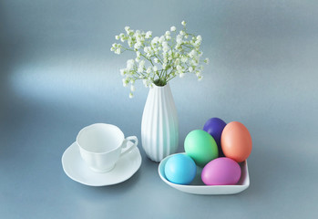A white porcelain saucer with colored eggs, an elegant vase with a bouquet of fresh gypsophila, and a coffee Cup with a saucer next to it on a gray background. Selective focus. Easter Breakfast. 