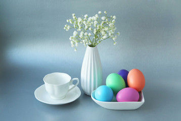 Beautiful Easter still life.It is Easter morning.A white porcelain vase with a gypsophila, white Cup and saucer,and plate with colored Easter eggs next to it on a gray background.Selective focus