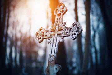 An old rusty beautiful crooked cross stands in the middle of a gloomy forest illuminated by the rays of the setting sun.