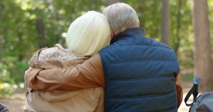 Back view of old Caucasian married couple resting in forest. Male and female senior tourists sitting on tree and hugging. Rear of senior man and woman embracing in wood. Hiking and tourism concept.