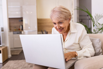 Portrait of senior woman sitting at table at home and working on her laptop. Older lady surfing the net from home while sitting on her sofa and using laptop computer