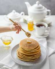 A children's hand pours honey on a stack of American pancakes. Fresh homemade sweet breakfast with tea. Bright food still life in a light key. Postcard to the day of pancakes.