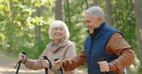 Senior Caucasian man and woman hiking with sticks in forest and talking. Happy old couple having conversation while walking with trekking poles in wood. Roaming and traveling concept. - Powered by Adobe