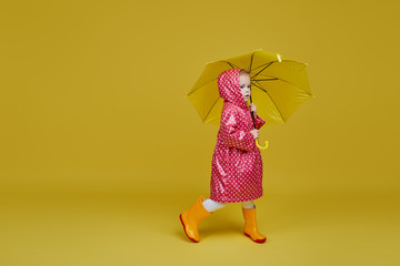 Cheerful child girl with yellow umbrella and red rain coat on colored yellow background. Copy space...