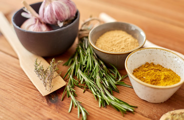 food, culinary and eating concept - bowls with different spices, rosemary, spatula and garlic on wooden table