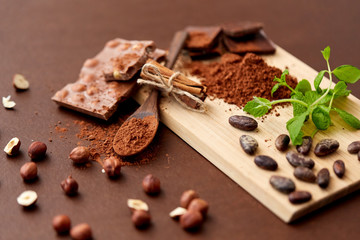 sweets, confectionery and culinary concept - chocolate with hazelnuts, cocoa beans, powder and cinnamon on wooden board with spoon on brown background