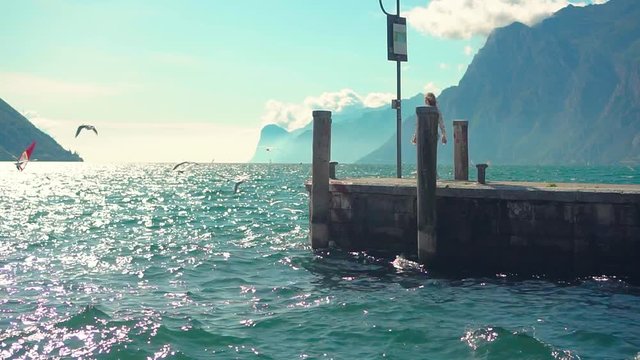 Girl in white short dress walks along pier, hair fly in wind, turned away enjoy divine view nature waves turquoise water. Seagulls fly over lake Lago di Garda sailboat floats. olt town Rina Del Italy