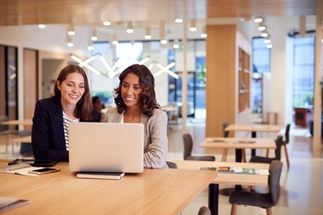 Two Businesswomen With Laptop At Desk In Open Plan Office Collaborating On Project Together