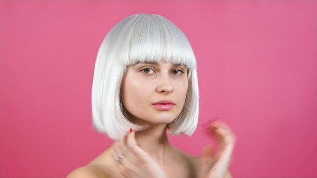 young beautiful girl in a blond bob wig, close up, posing in studio on a pink background. straightens her hair, preens. looks like in the mirror