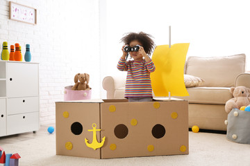 Cute African American child playing with cardboard ship and binoculars at home