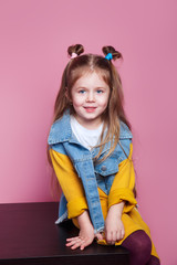 Beautiful stylish little girl in jeans clothes sitting and smiling on pink background