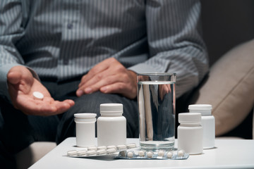 Overworked businessman sitting on the sofa and taking medication. Pills and glass of water near him.