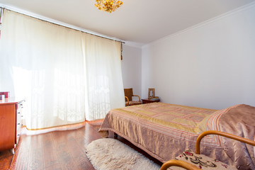 A large bedroom with a mahogany bed, wicker chairs, and an elegant dressing table with a semi-circular mirror. The bed has a beige blanket in squares. mahogany parquet