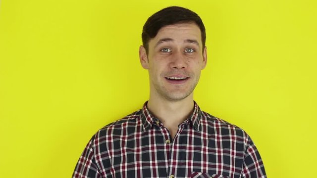 Young handsome guy in a plaid shirt, says goodbye and waves his hand .Portrait on a yellow background.
