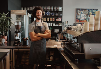 Successful male business owner behind the counter of a coffee shop with folded hands smiling looking at camera