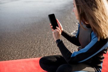 Portrait of a young relaxed surfer in wetsuit sitting with surfboard and smart phone on the beach. Water sport and leisure activity concept