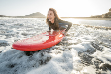 Young woman in wetsuit catching water flow on the surfboard, surfing on the wavy ocean during a sunset. Water sports and active lifestyle concept - obrazy, fototapety, plakaty