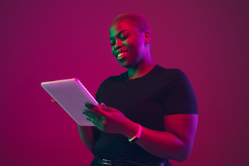 Using tablet. African-american young woman's portrait on purple background. Beautiful model in black shirt. Concept of emotions, facial expression, sales, ad, inclusion, diversity. Copyspace.