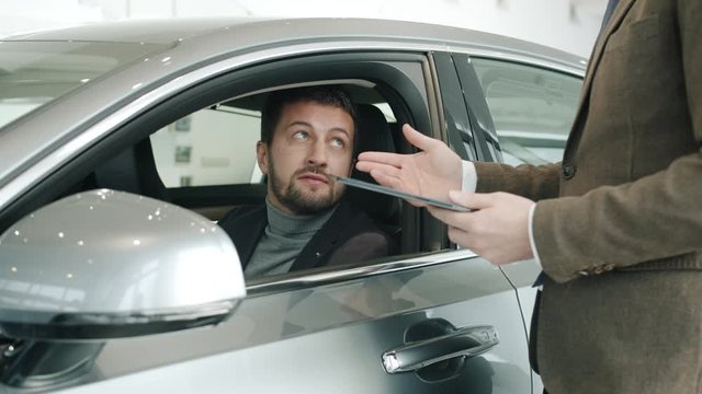Good-looking man buyer is talking to sales manager sitting in car touching shiny auto while salesman is discussing new model holding tablet computer.