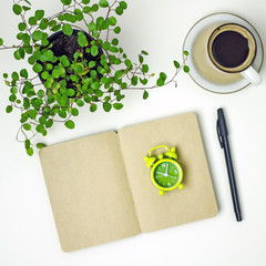  Coffee time. Top view on a white table with a notebook with dark sheets, pen, green alarm clock, indoor flower, cup of coffee. Time to drink coffee, coffee break, Working day concept, flat lay