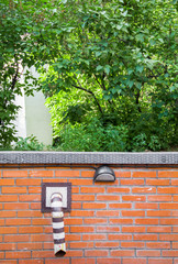 red brick wall with a striped black and white sewer tube against a background of bright green foliage on a tree